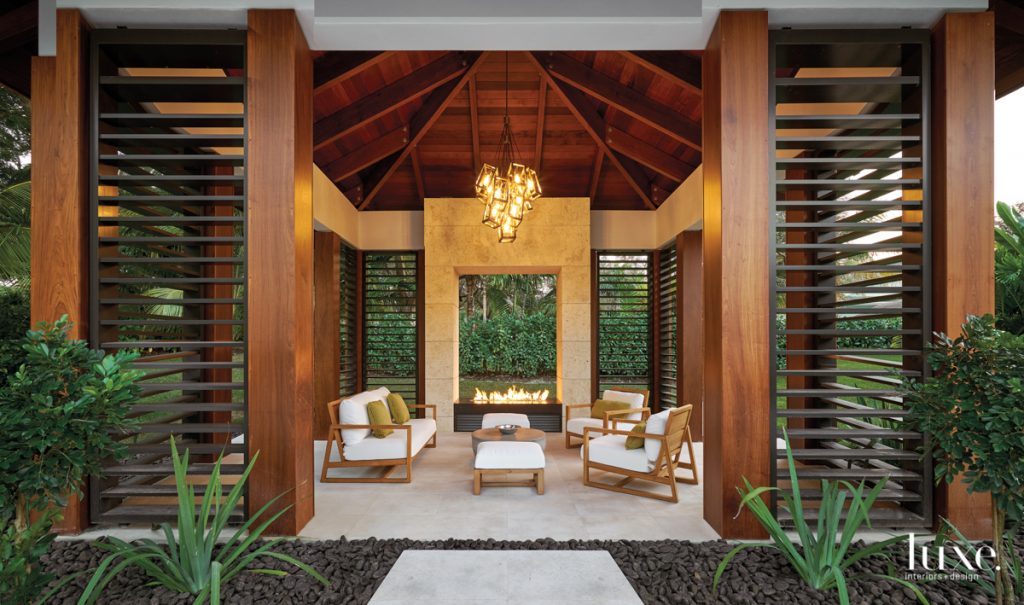 Beneath a Hinkley chandelier from Linear Lighting & Control, Summer Classics seating with pillows in a Romo fabric gather around an RH coffee table in a poolside pavilion at a Pinecrest home by interior designer Ivette Arango. An open-concept fire pit occupies a surround made of Opustone's filled coral and lava stone.