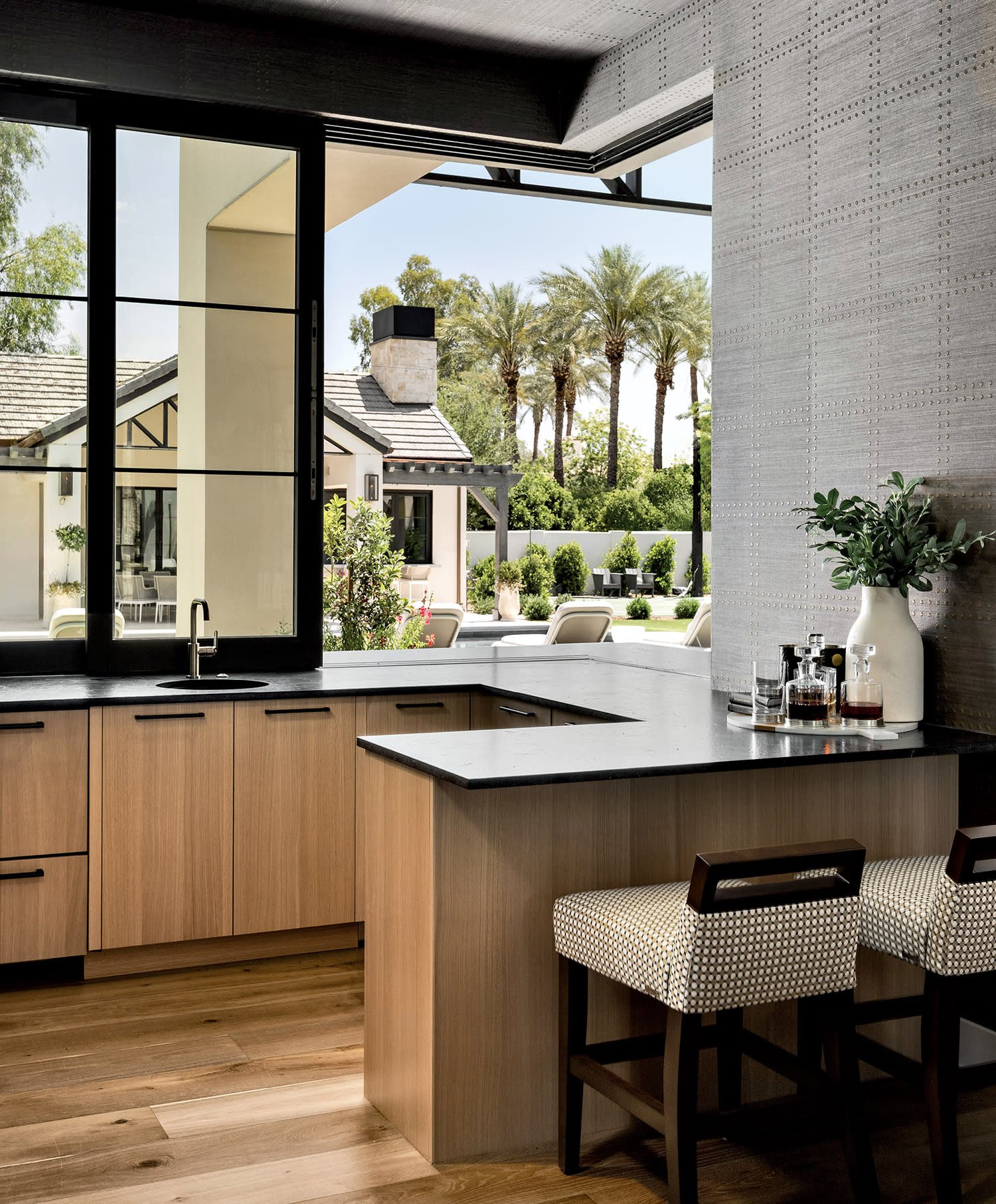 Modern kitchen with light wood cabinets and a black countertop. Two patterned chairs sit at the counter.