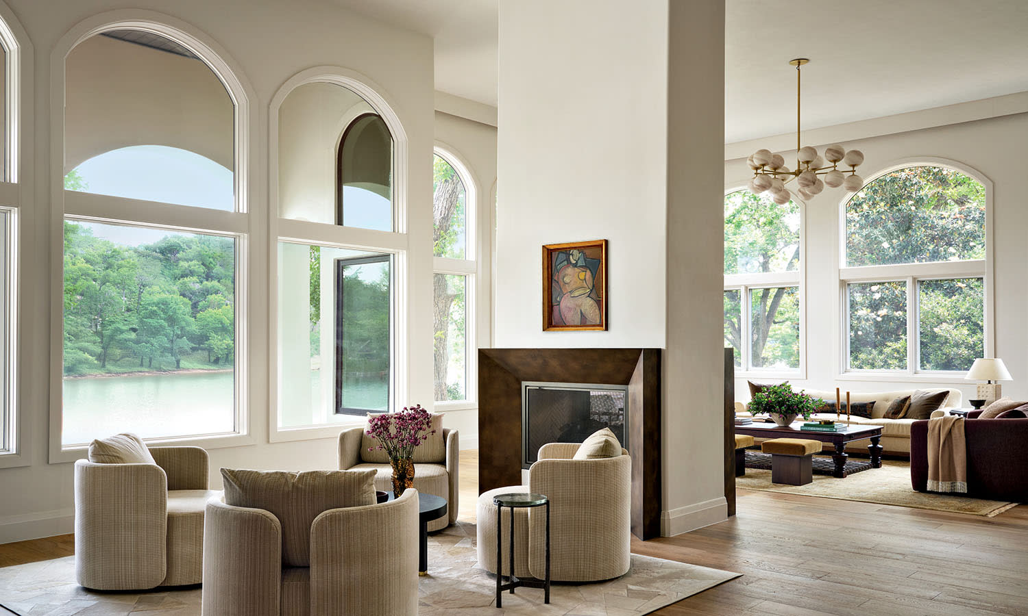 Spacious living room with large arched windows, beige armchairs around a small table, and a stylish fireplace. Bright, serene atmosphere with tree views.
