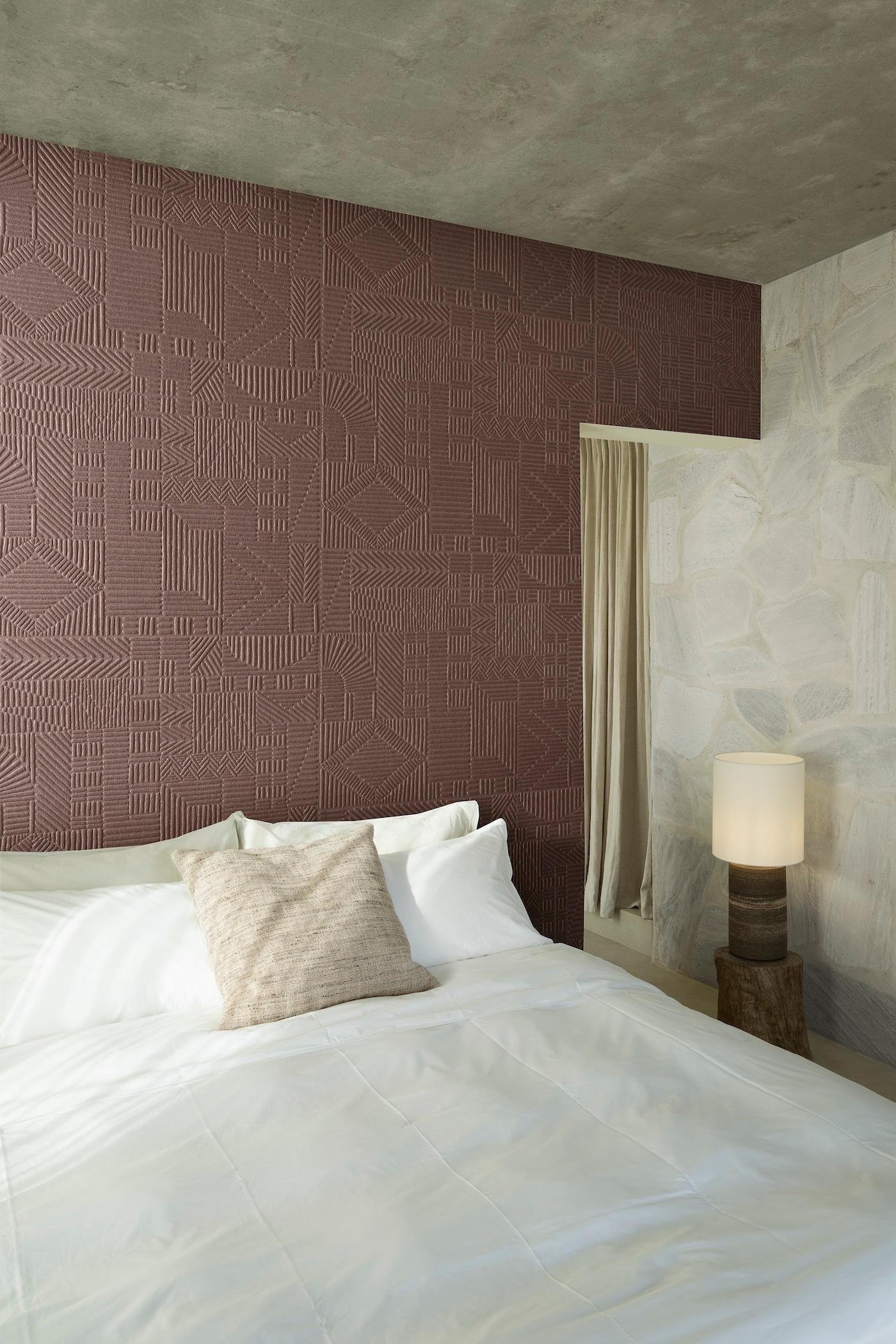Modern bedroom with a textured maroon accent wall, white stone walls, and a bed with crisp white bedding and a beige pillow. Warm lamp on log base adds coziness.