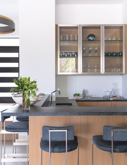 A modern kitchen with a counter, bar-style seating, glass-front cabinets, and a sink. There is a bowl of green foliage on the counter and various glassware on display in the cabinets.