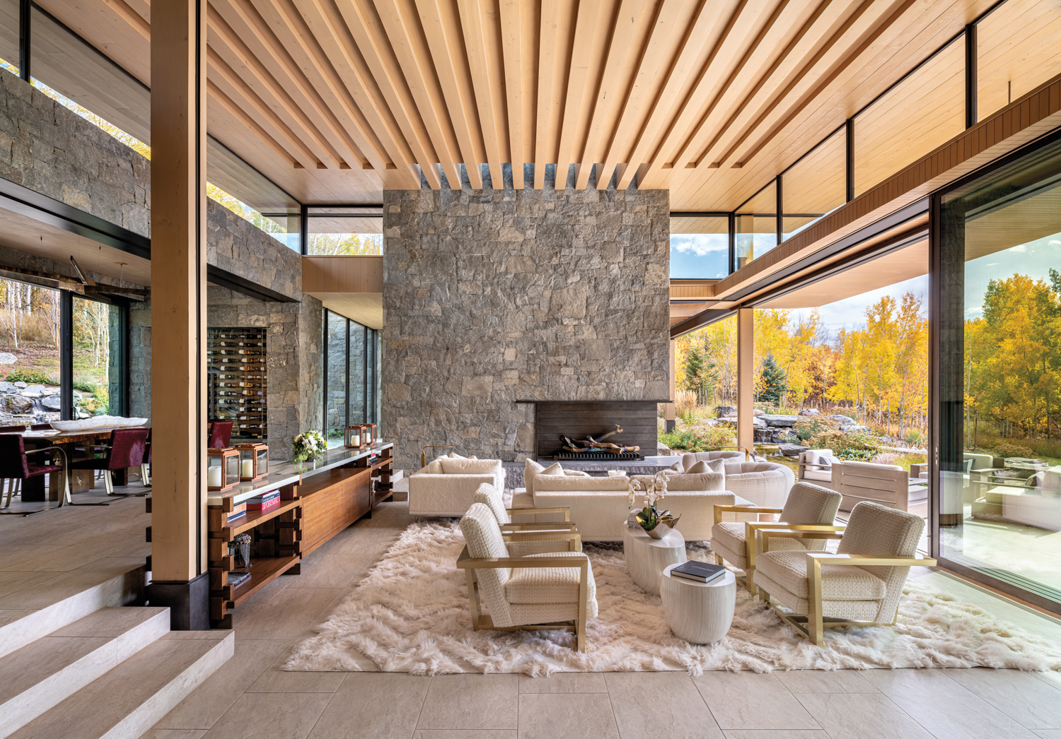 Great room with granite fireplace surround, slatted-wood ceilings, window walls with landscape views, and furnishings in soft, white materials
