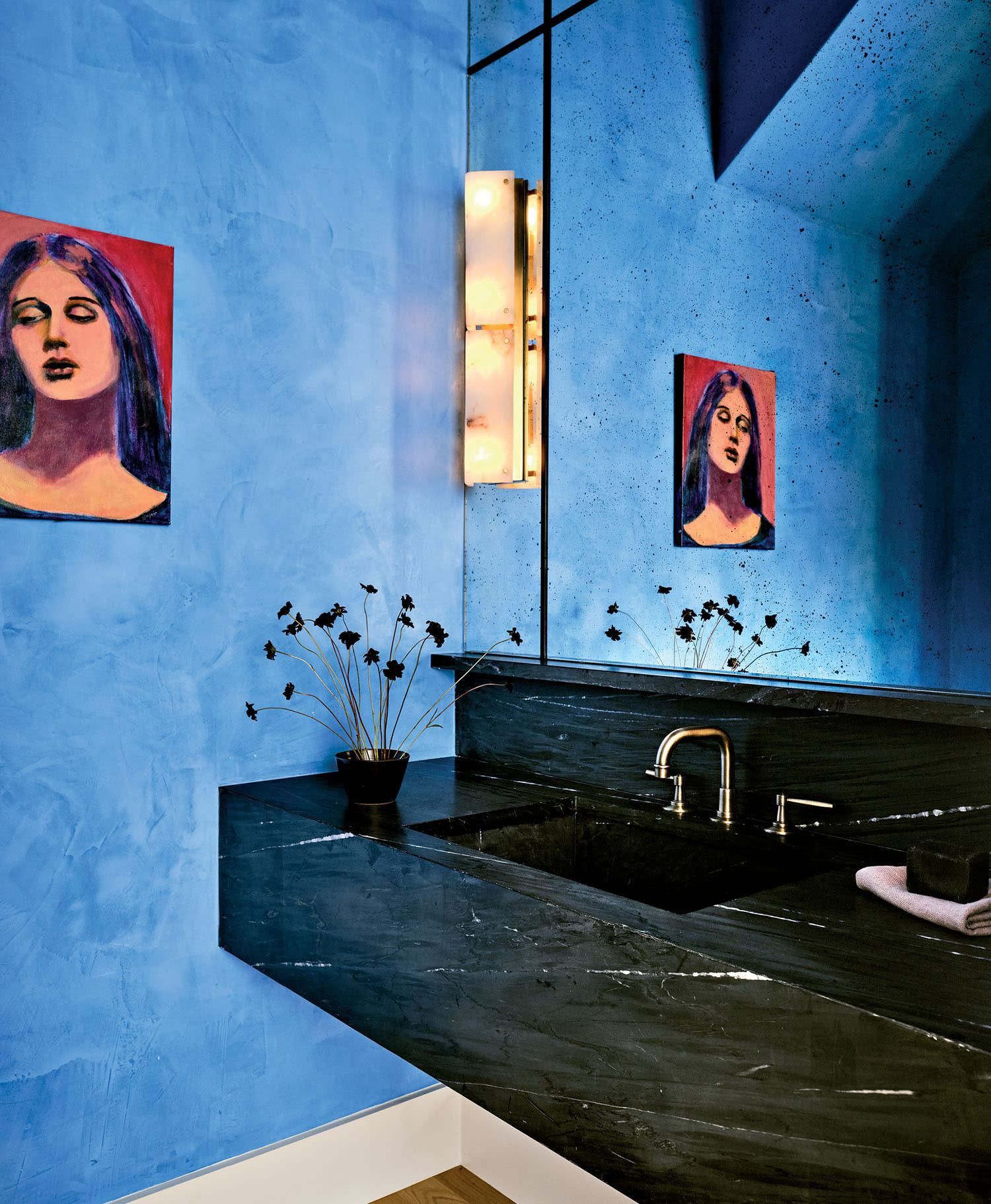 Modern bathroom with vibrant blue walls and a large mirror reflecting a painting of a woman. Black marble sink with minimalist decor and ambient lighting.