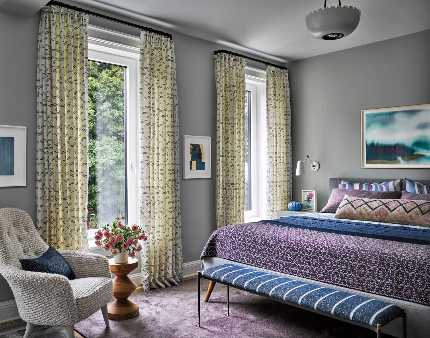 Cozy bedroom with gray walls, colorful bed linens, and patterned curtains. A plush chair and flowers add warmth. Sunlight filters through large windows.