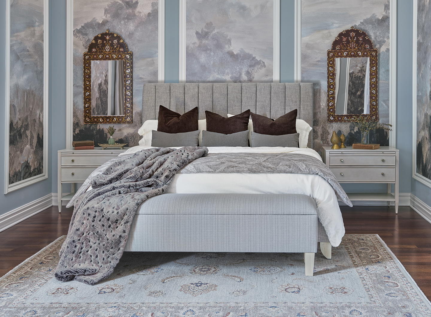 Elegant bedroom with upholstered gray bed, layered pillows, fur blanket, mirrored nightstands, ornate wall mirrors, cloud mural wallpaper, hardwood floors, and patterned vintage-style area rug.