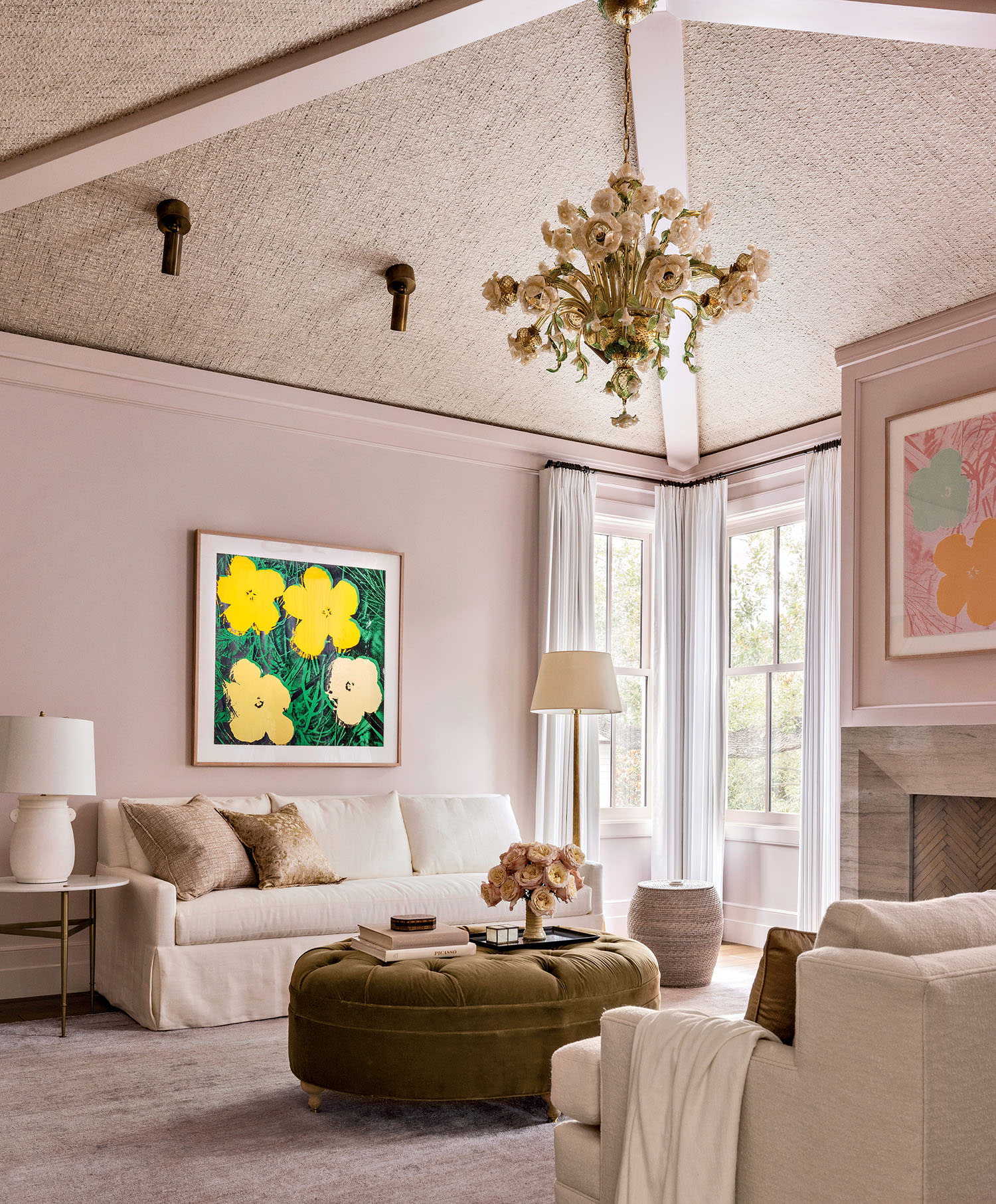 Elegant home office with a cozy sofa, brown ottoman, and floral paintings. A floral chandelier hangs above, next to large, light-filled windows.
