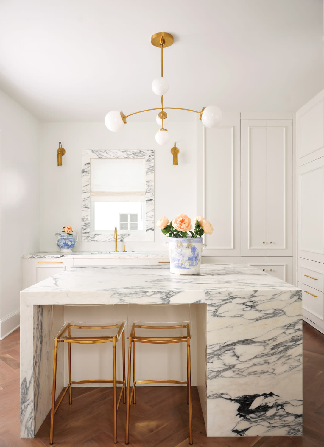 Modern kitchen with a marble island, gold stools, and a gold light fixture. Elegant decor includes a floral centerpiece and blue vase, creating a bright, stylish atmosphere.