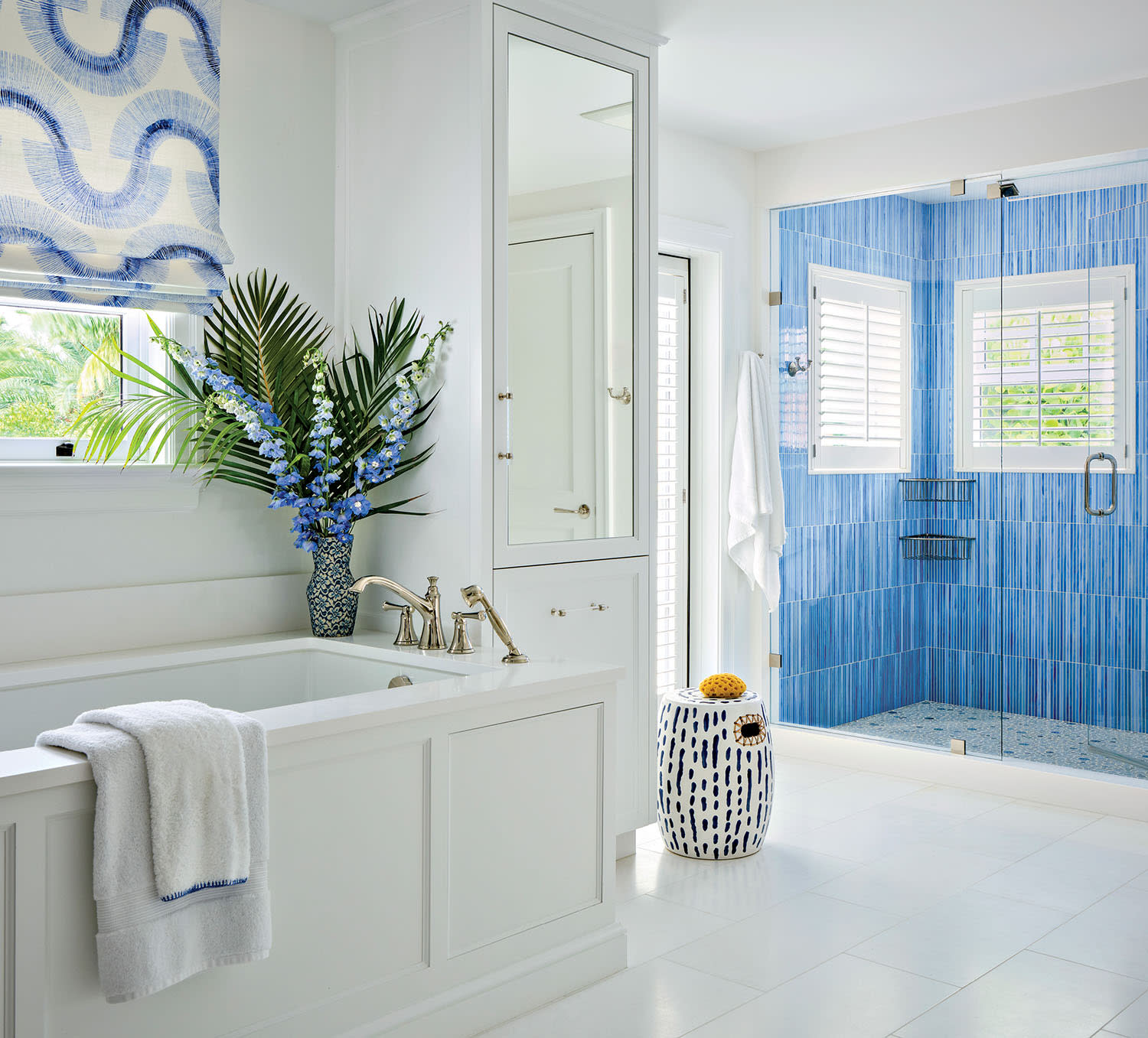 Elegant white bathroom with blue accents, featuring a large bathtub with gold fixtures, a blue-tiled shower, and lush greenery for a fresh feel.
