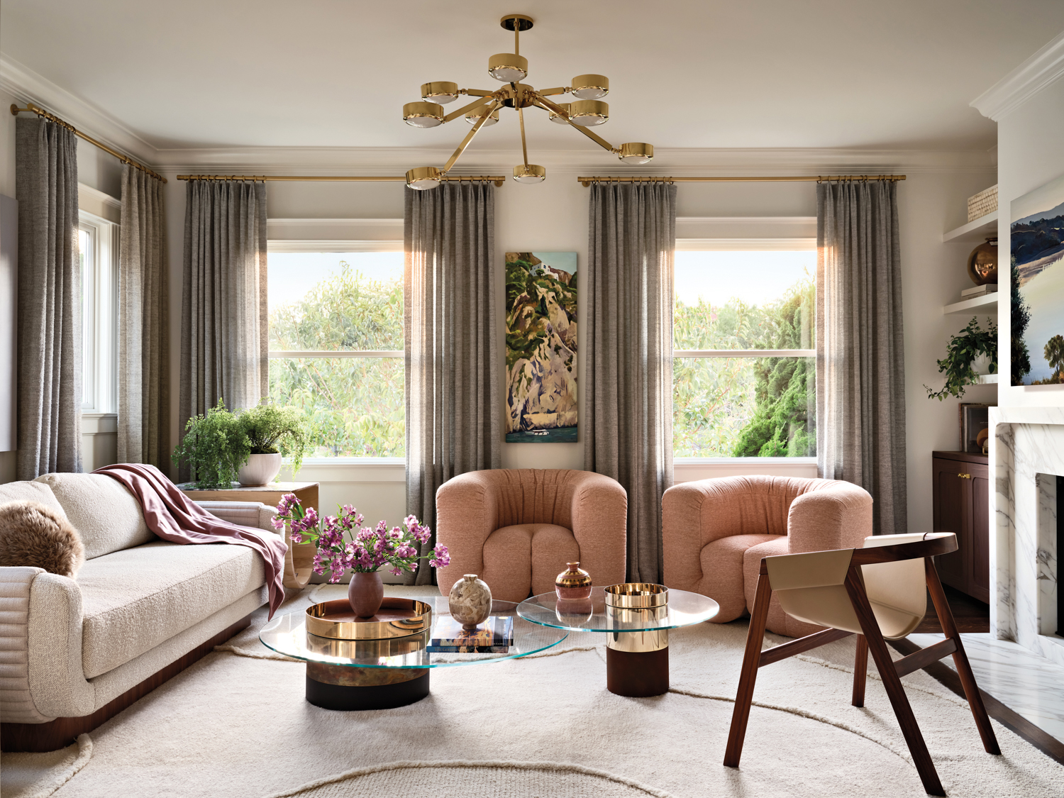 Elegant living room with a cream sofa, two pink armchairs, modern glass coffee table, and gold chandelier. Soft gray curtains frame large windows.
