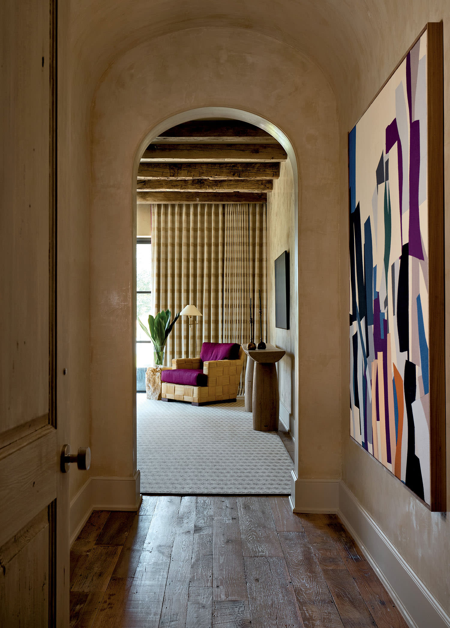A wooden-floored hallway leads to a cozy room with a yellow and purple chair. An abstract painting adorns the right wall, adding a modern touch.