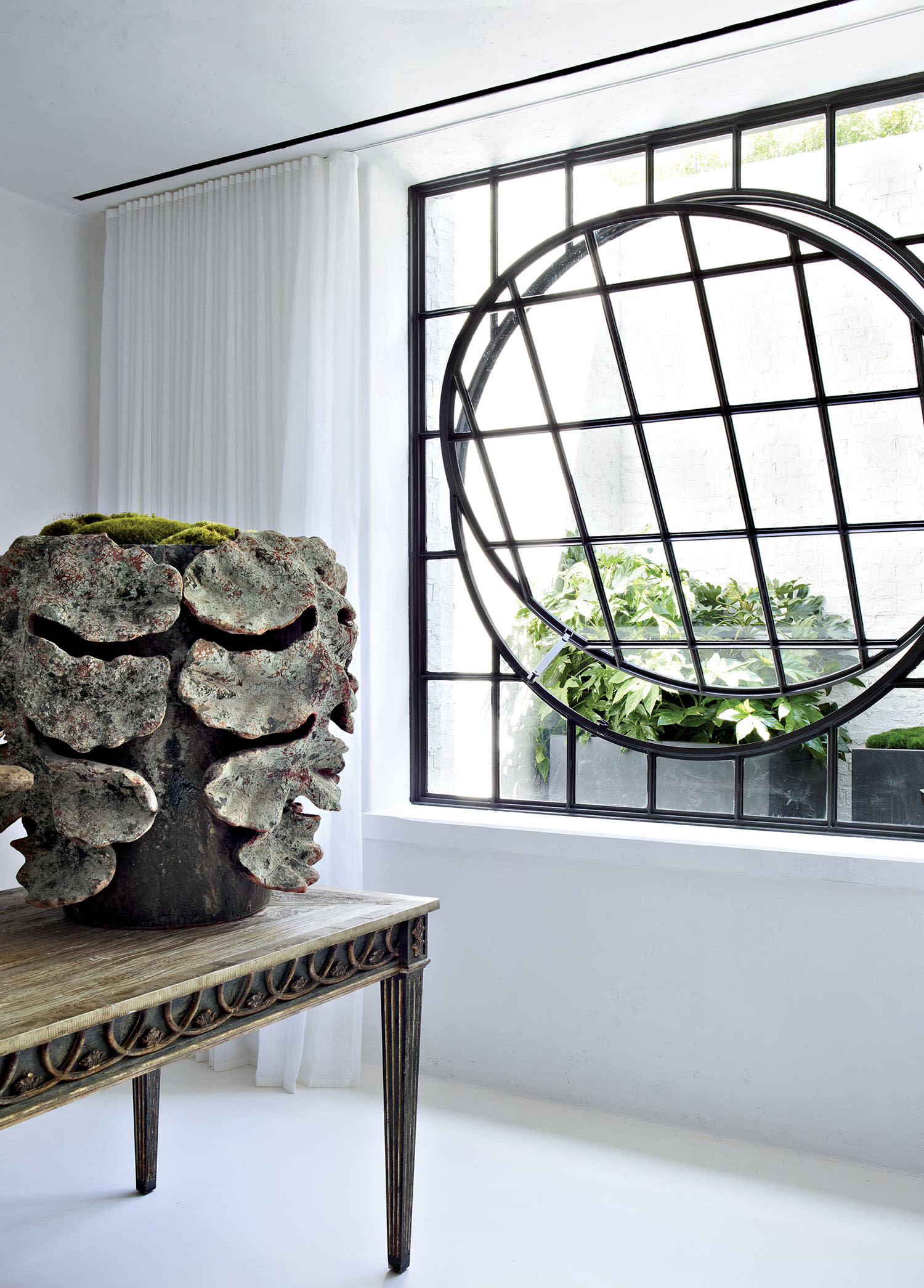 A wood table with plant sits in a white room with a window with circular frame partially open.