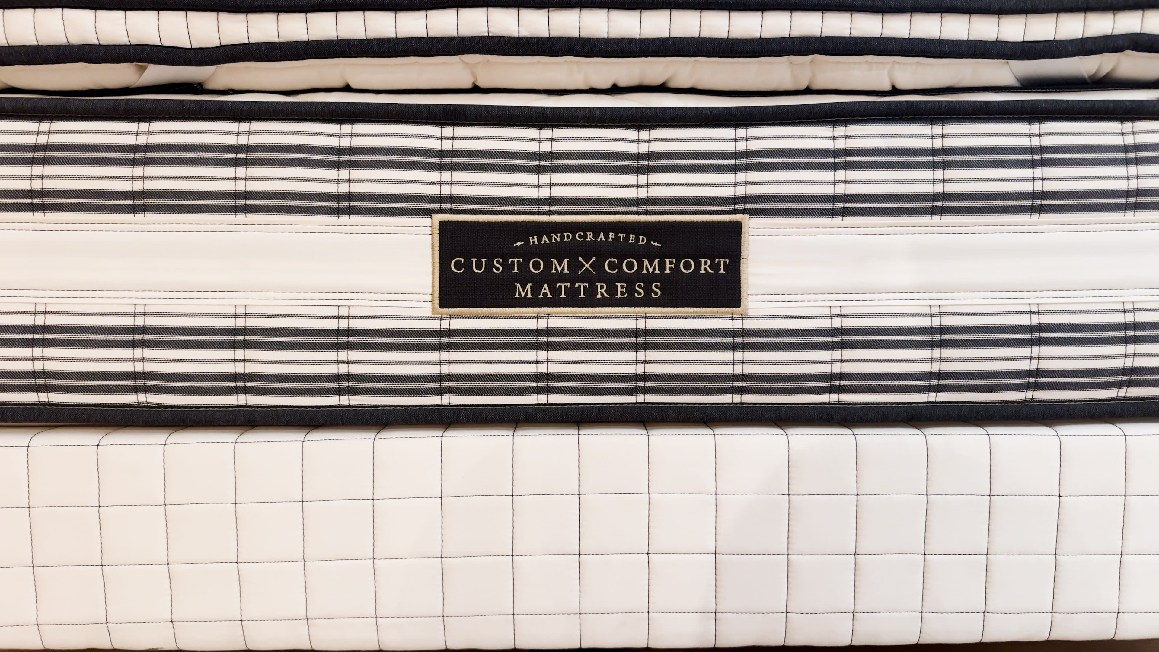Close-up of a mattress with striped and checkered patterns. A label in the center reads "Handcrafted Custom Comfort Mattress"