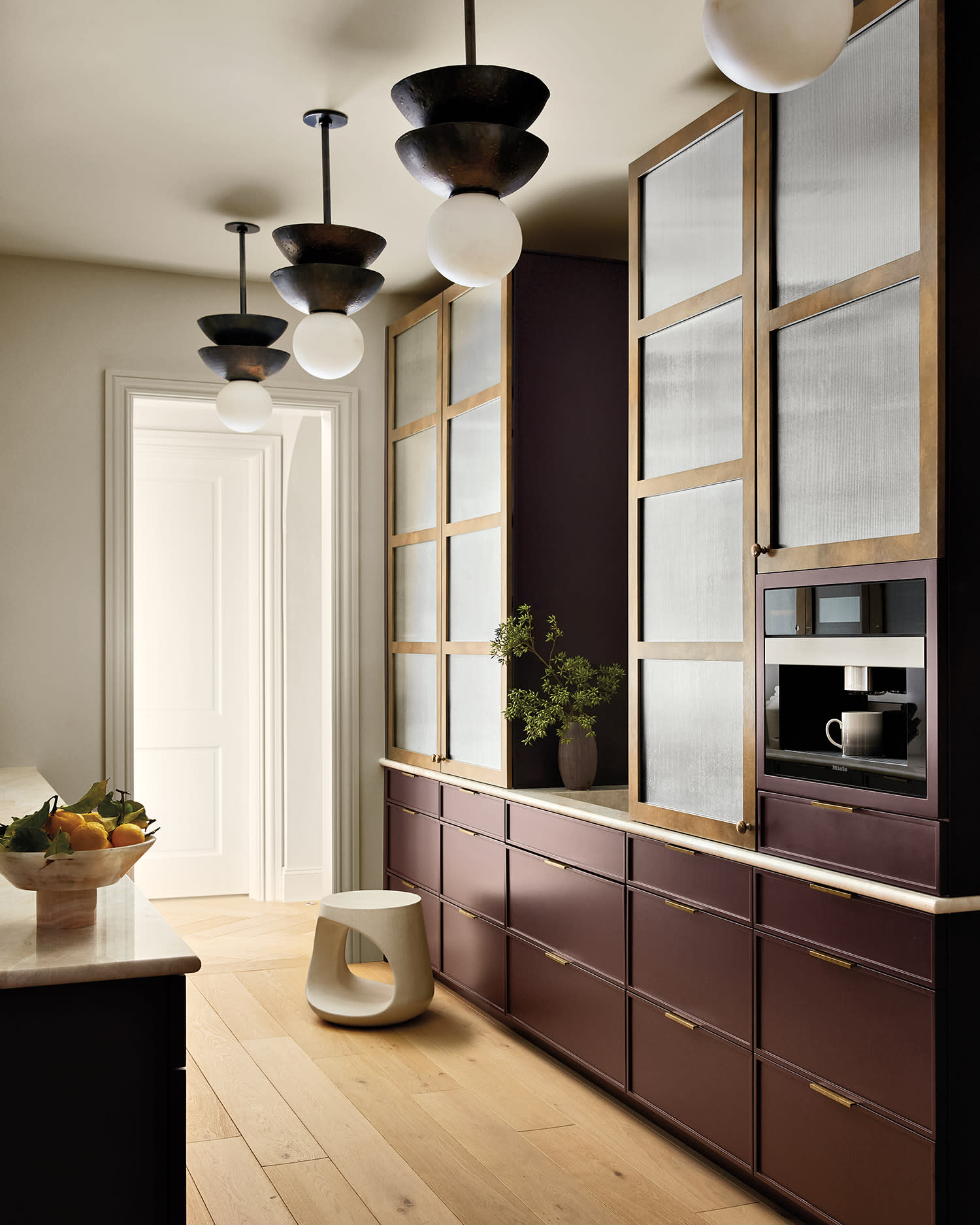 Kitchen pantry area with tall wood‑framed cabinets, brass‑glass pendant lights, and stool.