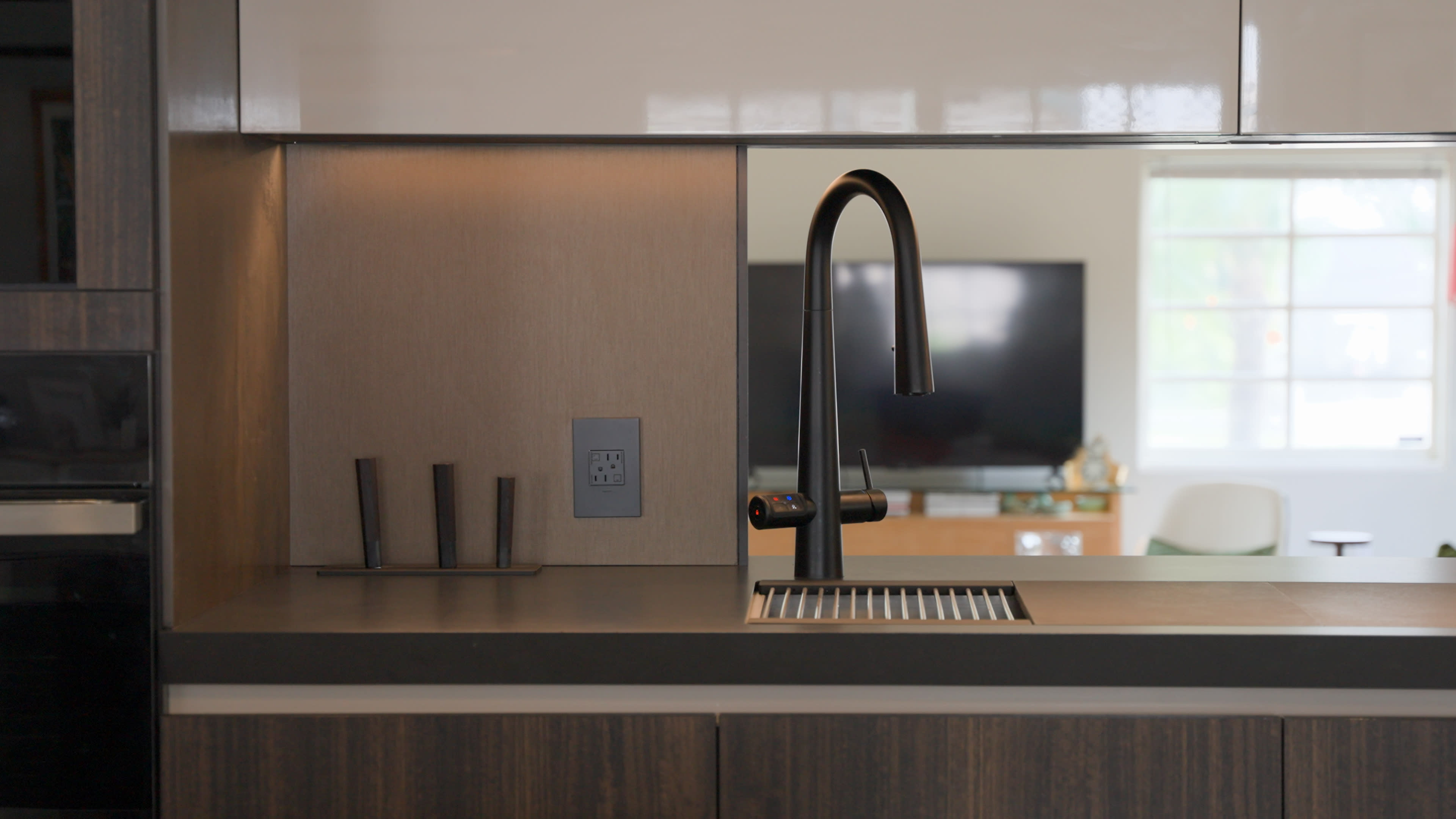 Modern kitchen interior with a sleek black faucet and dark countertop. Mood is minimalist and elegant. A TV and a window are visible in the background.