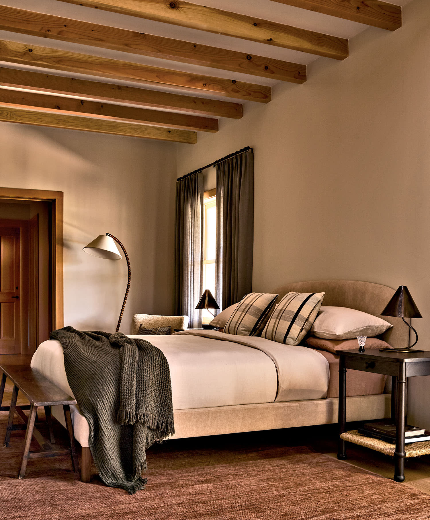 Cozy bedroom with exposed wooden beams, a beige bed with plaid pillows, and a knitted blanket. Soft lighting and neutral tones create a warm ambiance.