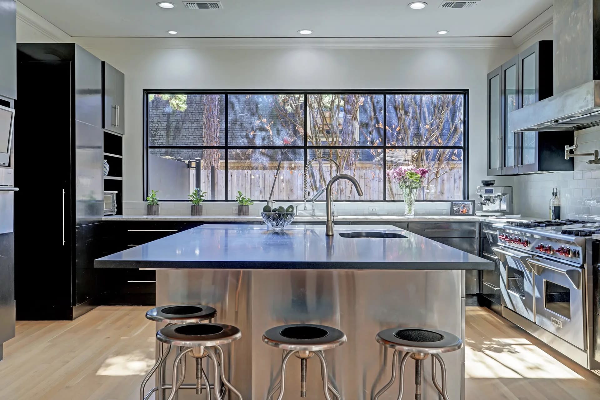 Modern kitchen with a large island countertop, three metal bar stools, stainless steel appliances, and large windows with a garden view, creating a sleek and bright atmosphere.