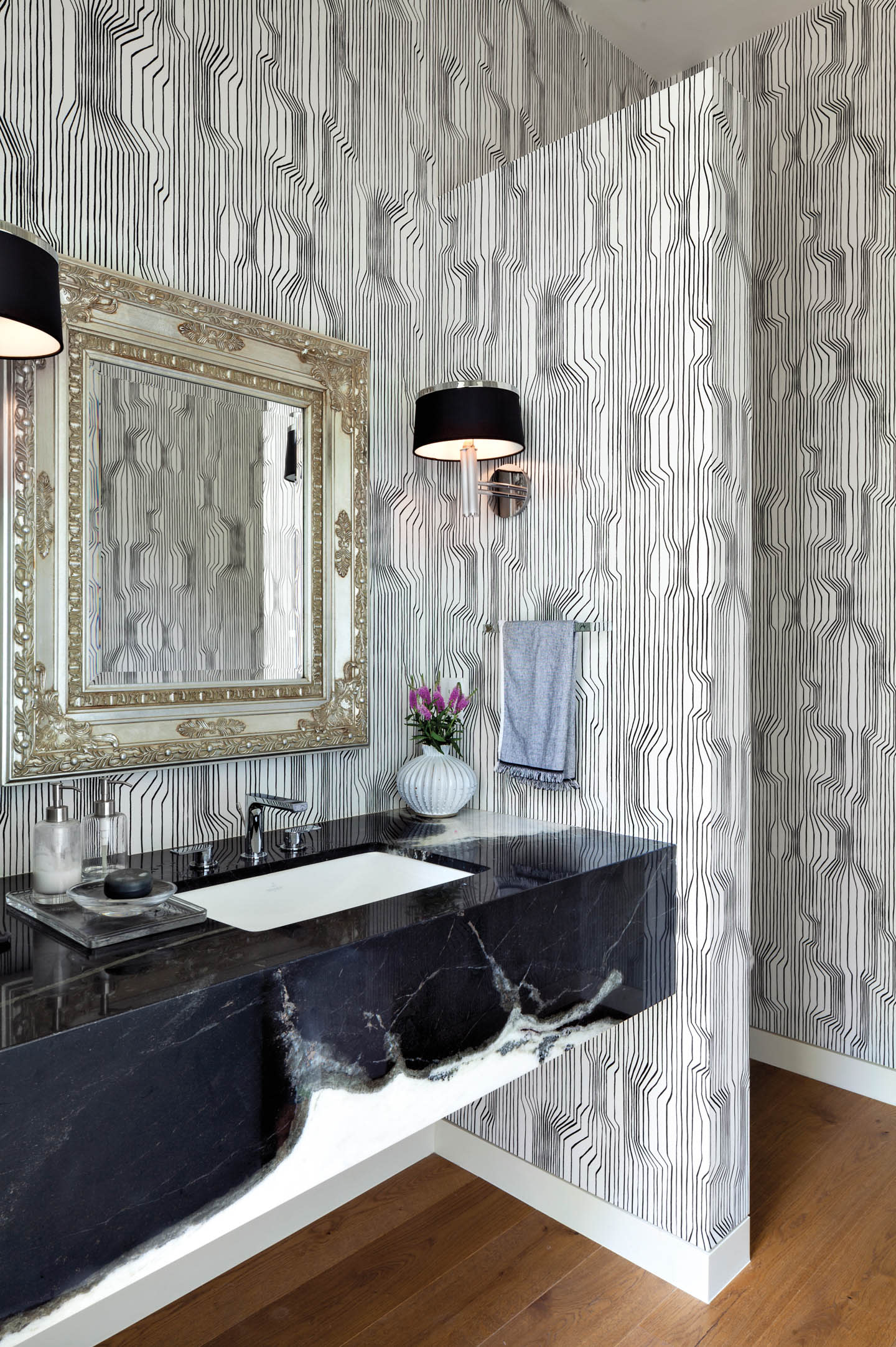 Black-and-white powder room with abstract line wallpaper, ornate mirror, black marble vanity, and wood floors.