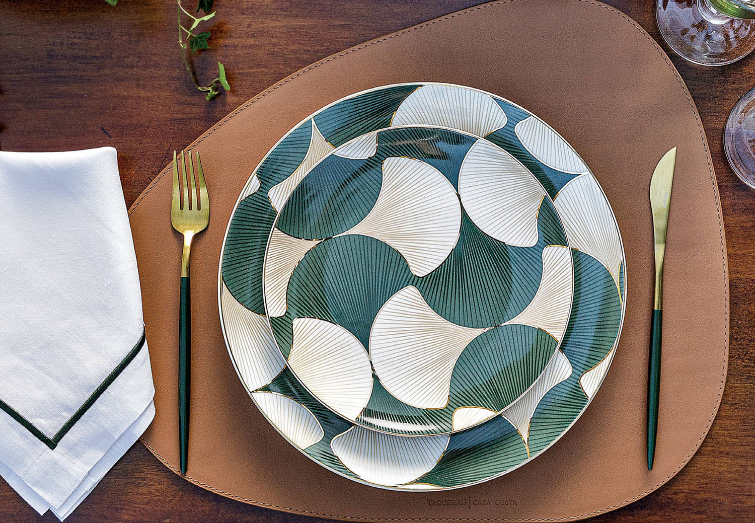A place setting with green-and-white patterned plates, gold-accented flatware and cloth napkin.