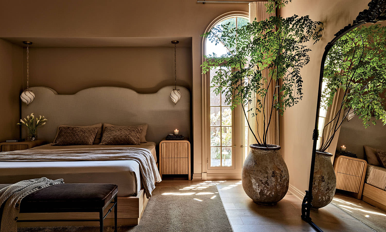 A bedroom in brown earth-toned hues with a bed, a mirror along the wall and a large plant