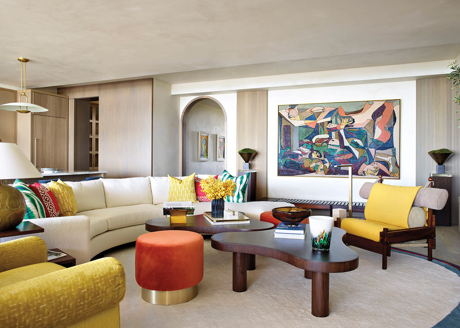 Stylish living room with a cream sectional sofa adorned with colorful pillows, abstract artwork, wooden coffee table, and vibrant orange and yellow accents.