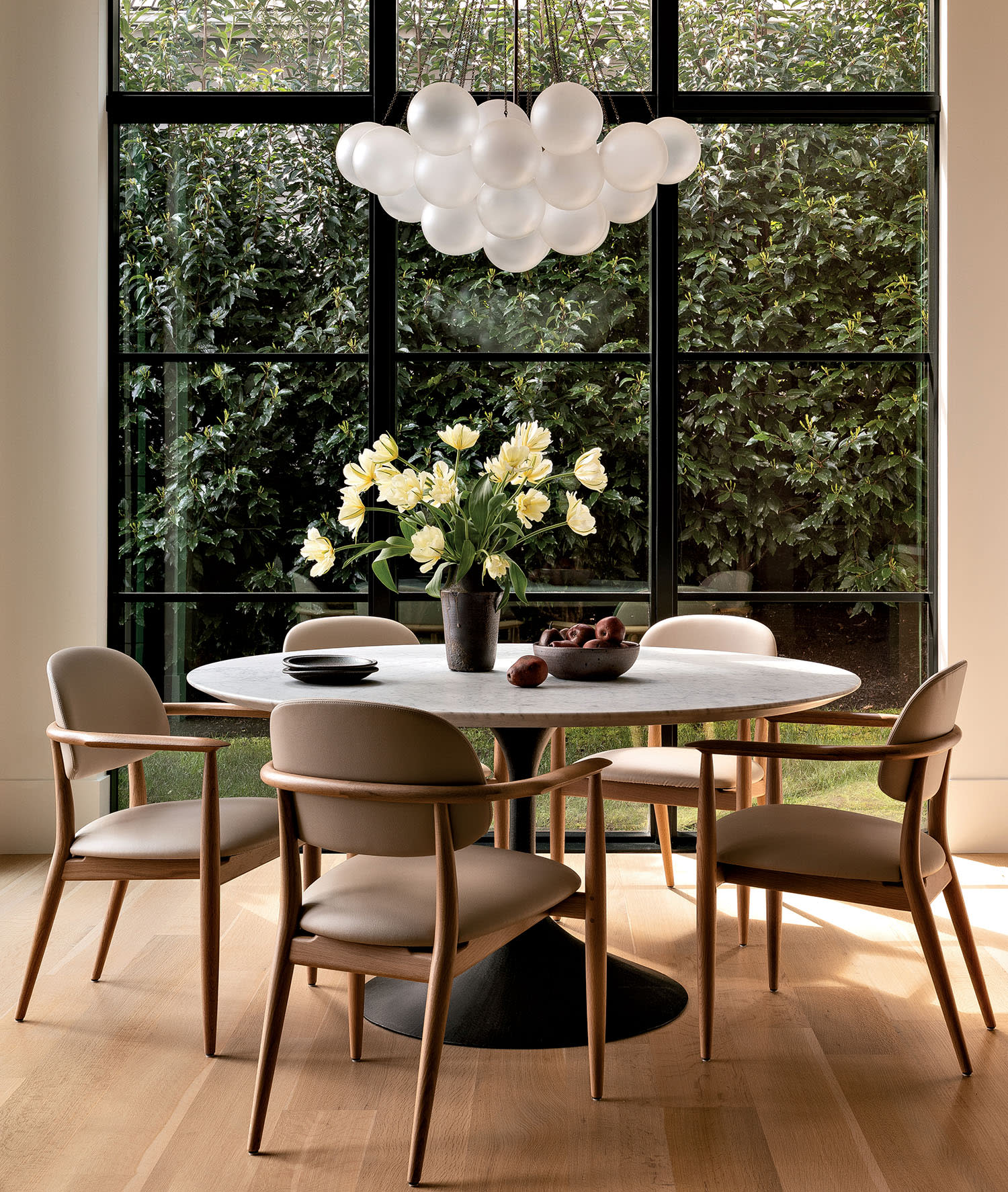 A modern dining room with a round table, beige chairs, and a centerpiece of white flowers. Large windows showcase lush greenery outside, and a contemporary chandelier hangs above.