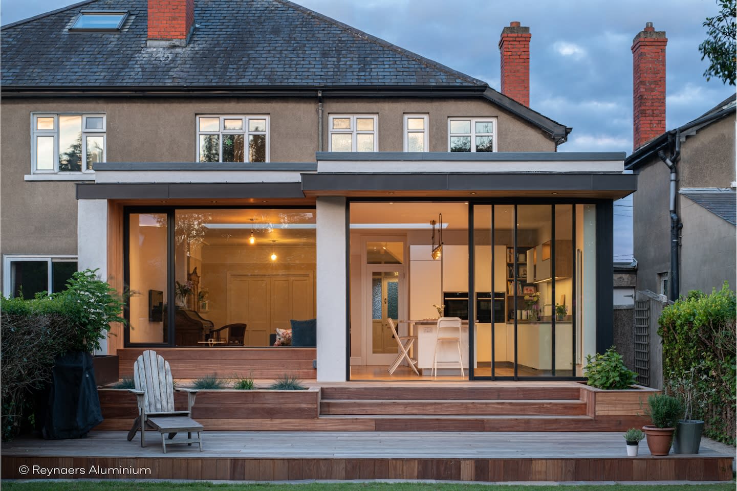 A modern home extension with large glass sliding doors, wooden patio and outdoor seating, blending indoor and outdoor spaces with warm lighting inside.