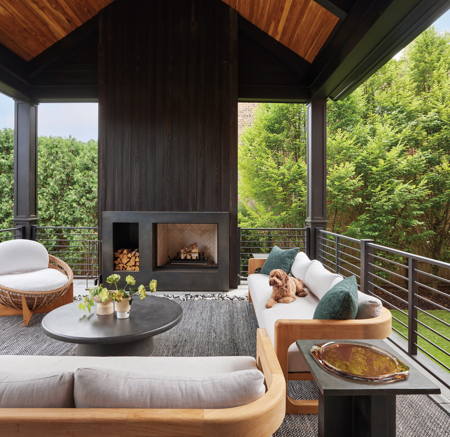 Stylish covered patio with a large fireplace, cozy seating, and a fluffy dog on the sofa. Surrounded by lush greenery, exuding a serene ambiance.