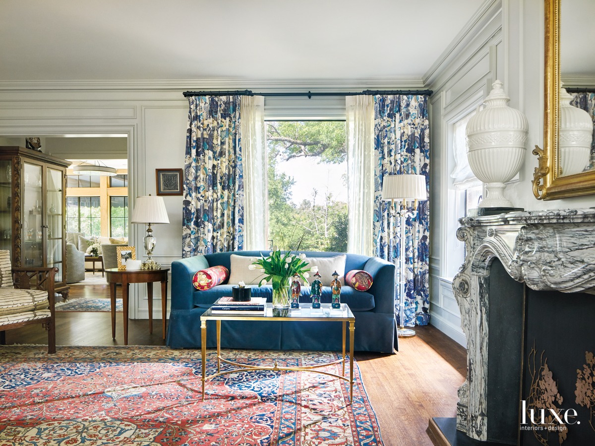 In the living room, a custom sofa wearing Schumacher velvet pairs with a coffee table from Global Views. McKinzie chose a Wind print from William & Wesley for the draperies and in a different colorway for the bolster pillows. The floor lamp and urn are from Decorative Crafts. The owners found the antique mantel and mirror on 1stdibs.