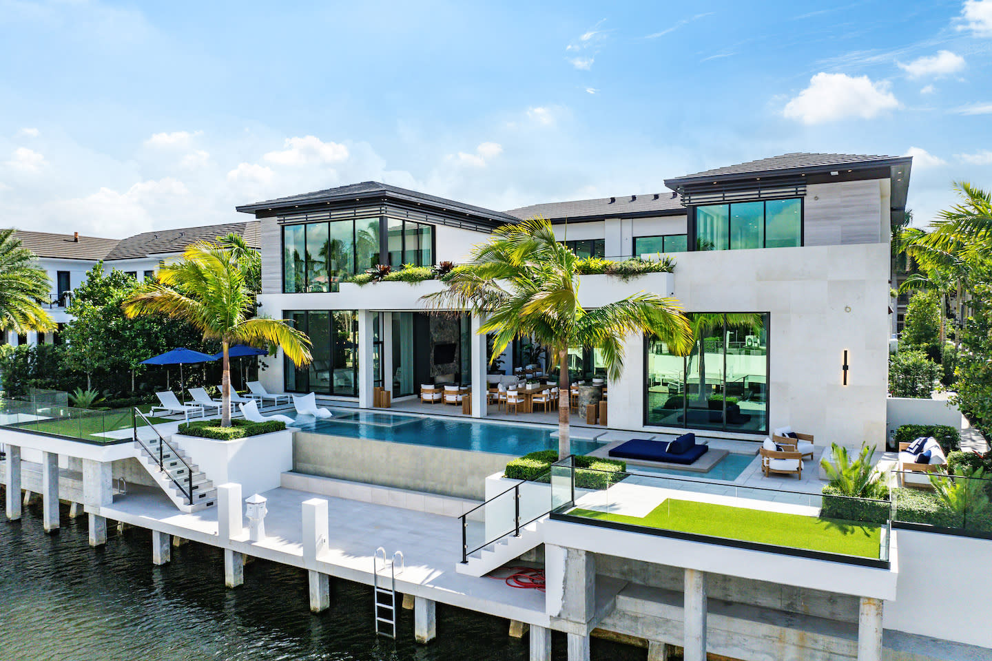 Modern waterfront estate with twin glass-walled wings and a sleek infinity pool that cantilevers above a private dock. Palm trees, rooftop greenery, and cushioned sun-lounging zones frame the bright white stucco and stone façade, while a ladder descends straight into the calm canal below beneath a clear blue sky.