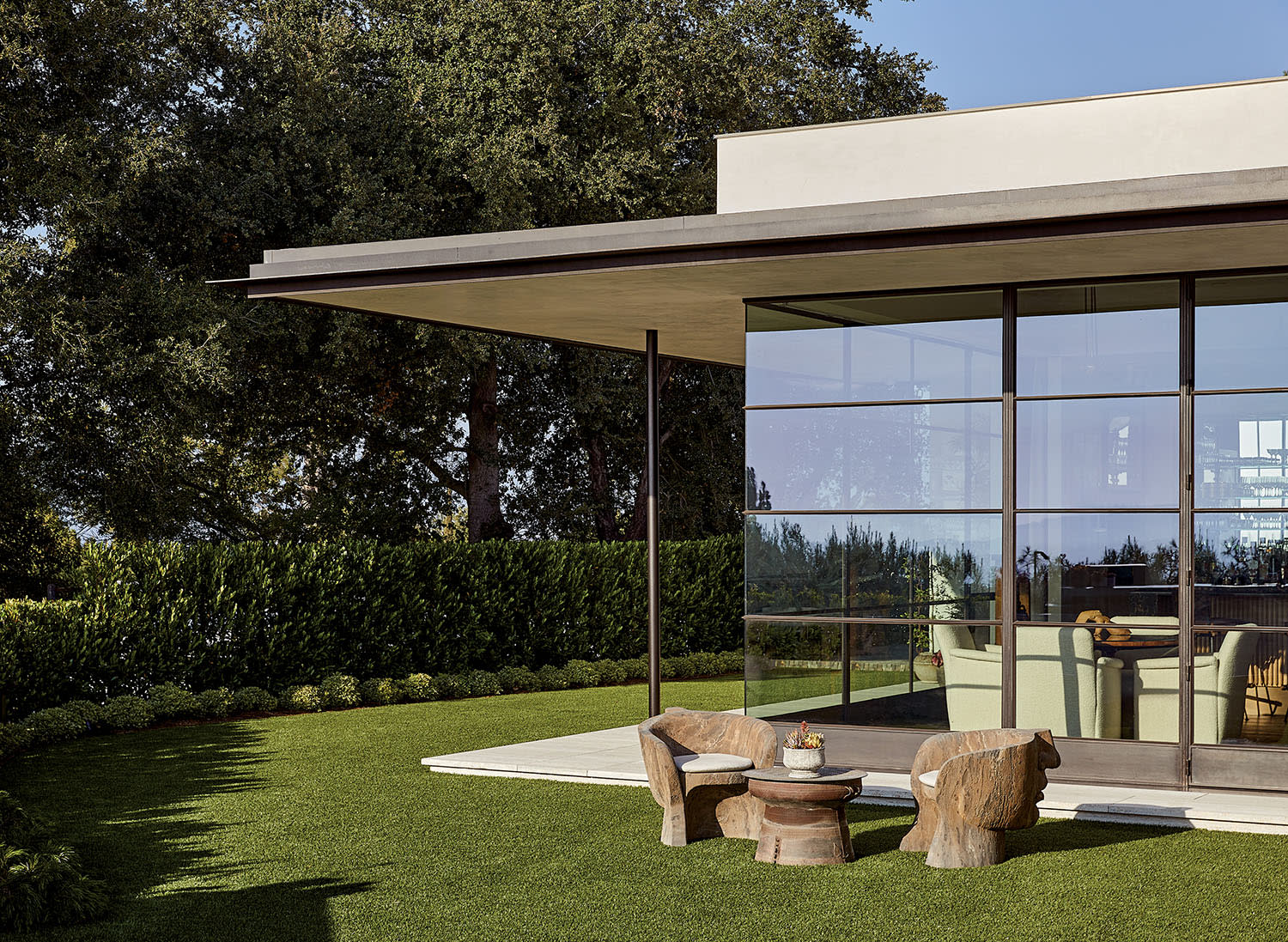 Modern glass-walled house with flat roof, surrounded by lush trees and greenery. Rustic chairs and table on a green lawn, creating a serene, stylish setting.