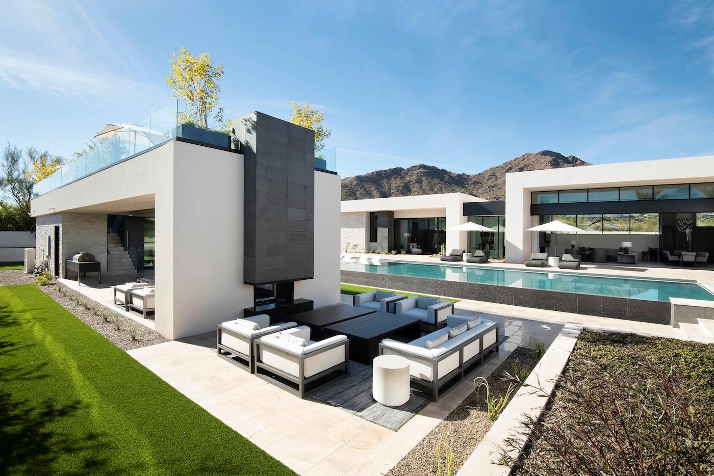 Modern luxury home with sleek white architecture, large swimming pool, outdoor seating, and mountain views under a clear blue sky. Tranquil atmosphere.