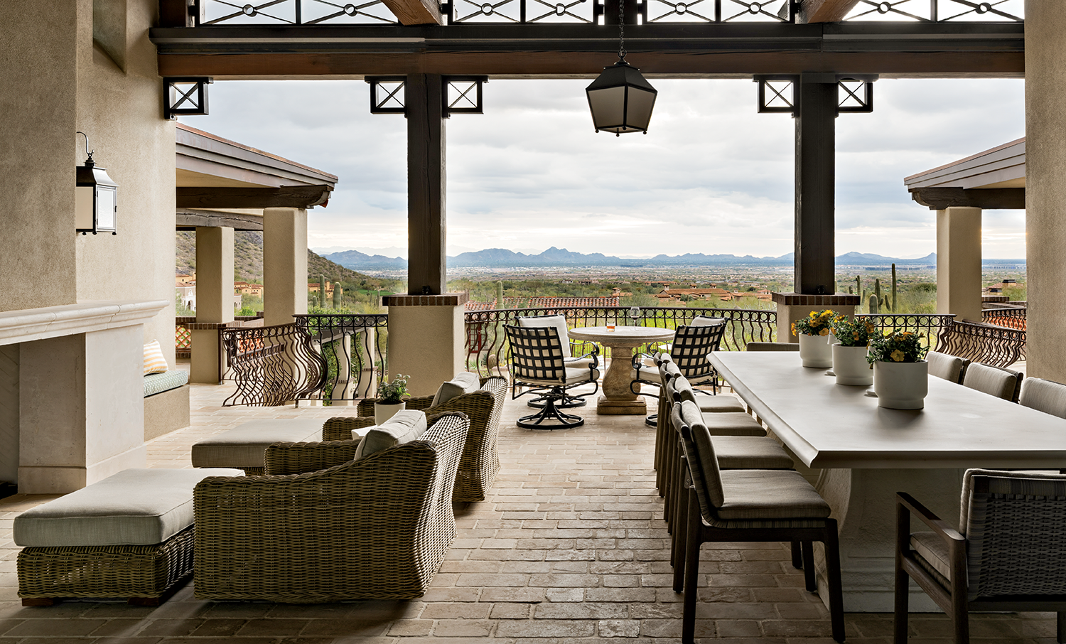 Rustic outdoor patio with wooden beams, wicker furniture, and a long dining table with potted plants