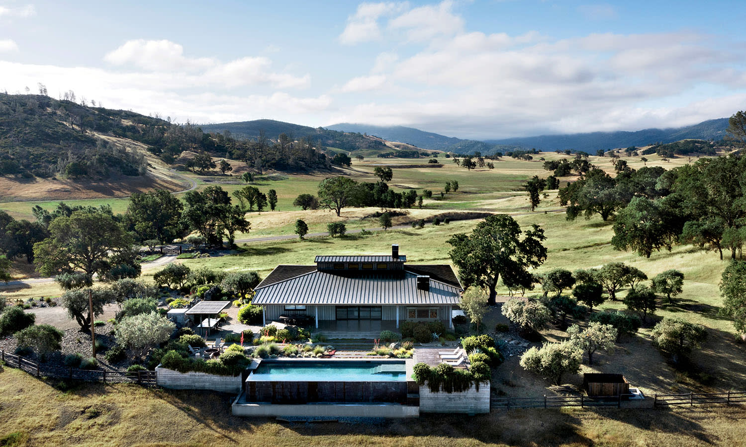 A modern house with a flat roof overlooking a scenic landscape. The property includes a swimming pool, surrounded by trees and rolling hills under a blue sky.