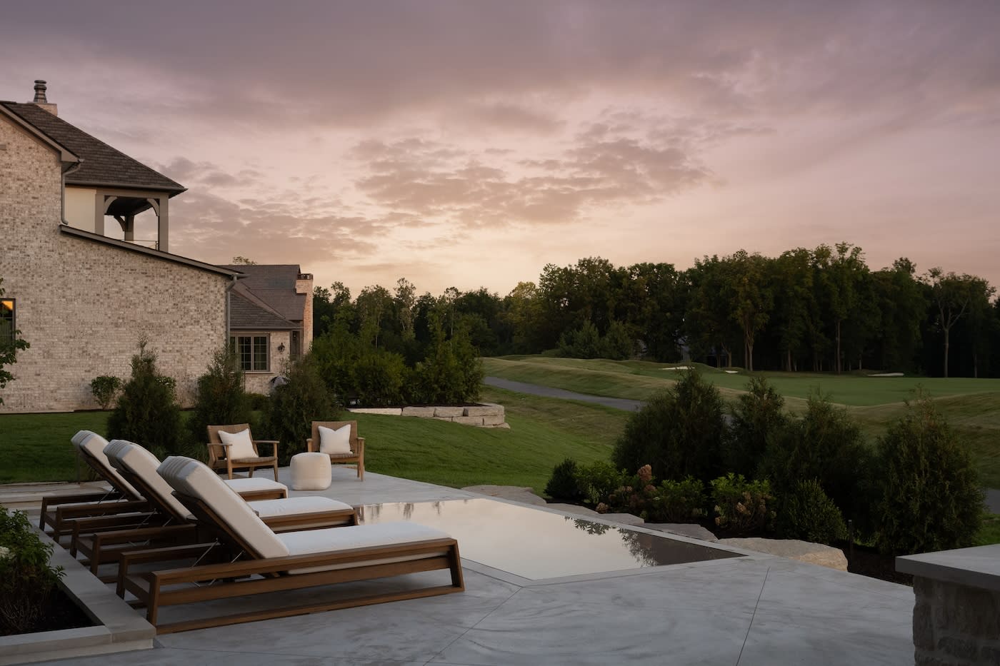 Poolside patio with wooden lounge chairs and cushions at sunset, adjacent to a large lawn and trees. The sky is pink and serene, enhancing tranquility.