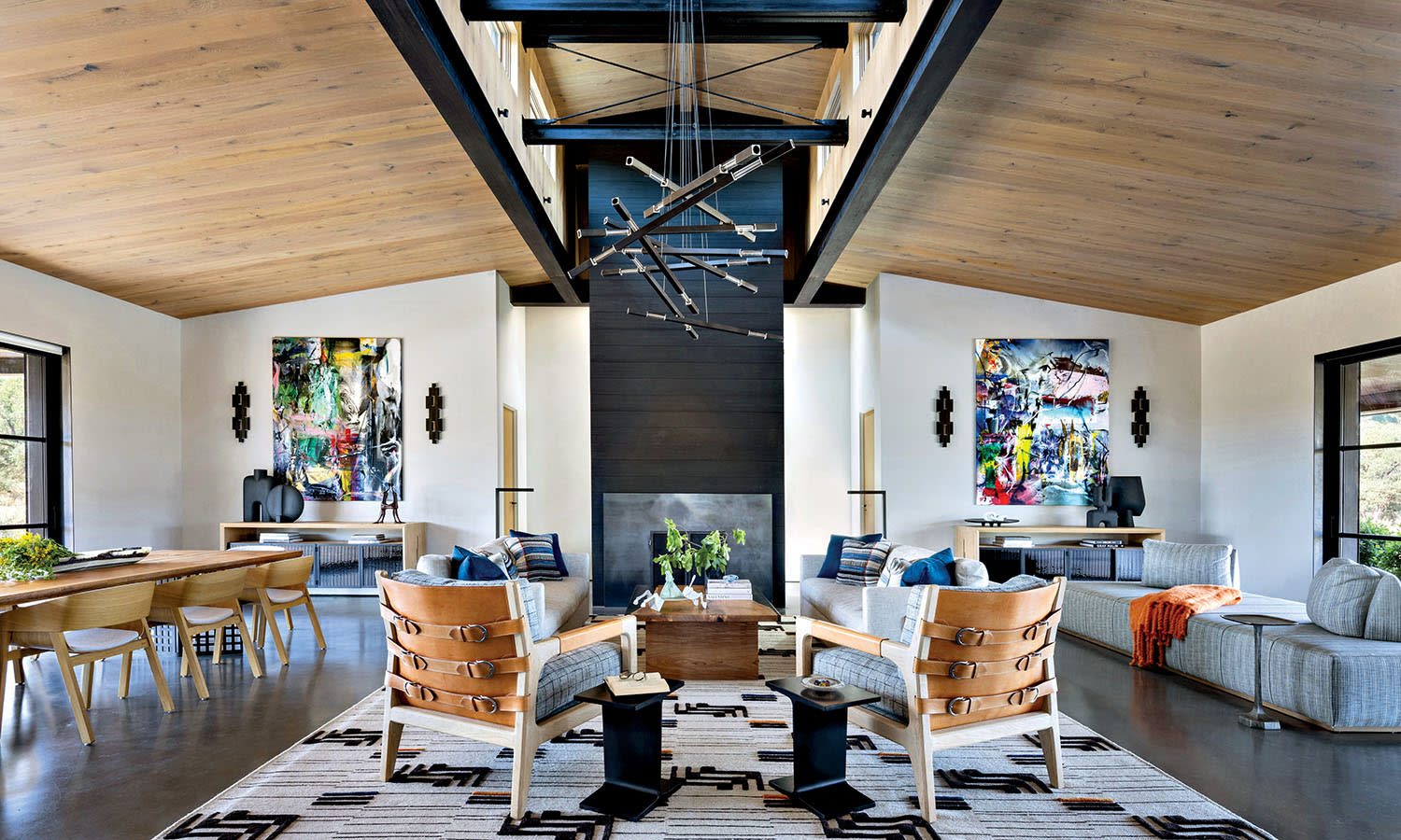 Spacious modern living room with high ceilings, wooden beams, and large windows. Features abstract art, cozy seating, a rustic rug, and a central geometric chandelier.