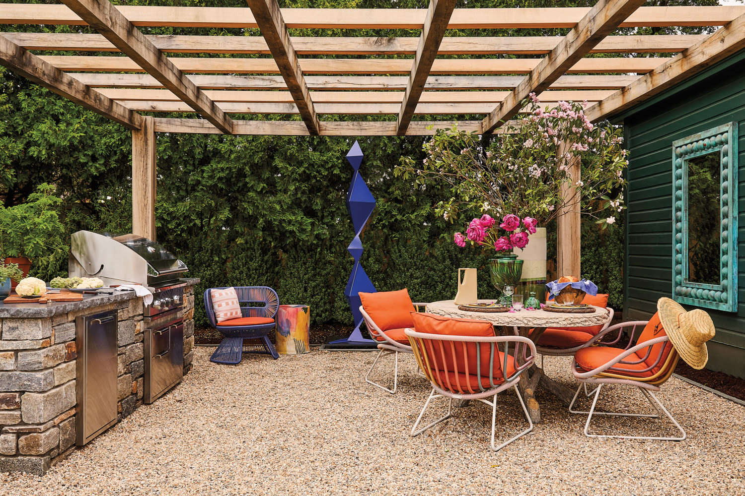 Outdoor patio with pergola and modern decor. Features a stone grill, colorful chairs, a round table, vibrant flowers, and a large plant. Relaxing vibe.