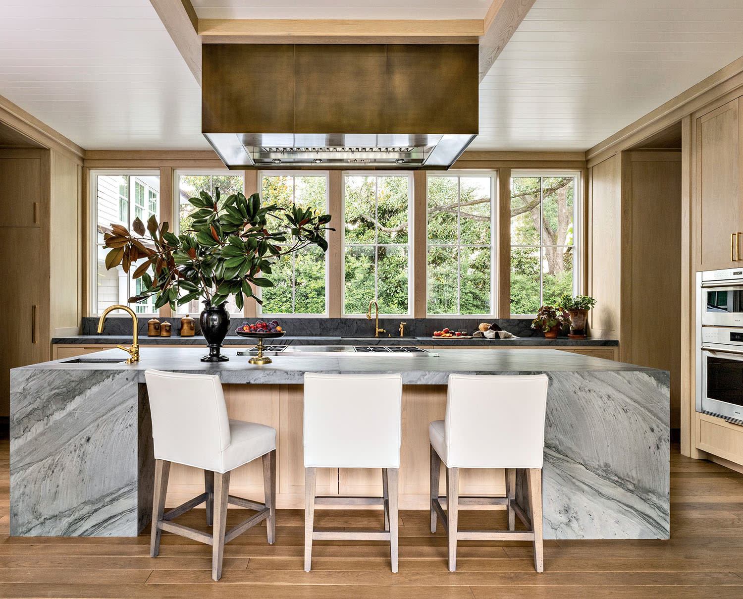 Sleek kitchen with marble island, three white chairs, and large windows revealing greenery outside. Brass fixtures add elegance and warmth.