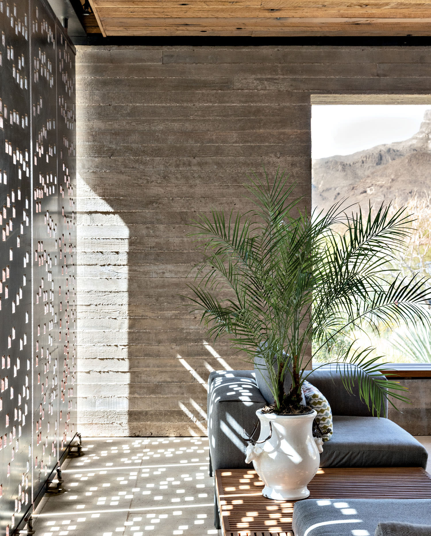 Light comes through perforated steel panels in a space with a windowed wall and low couches.