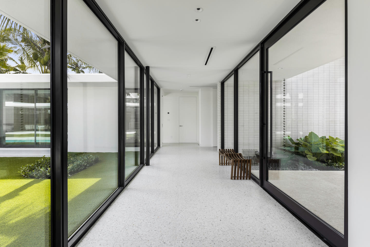 Minimalist glass-lined hallway with terrazzo flooring, flanked by black-framed floor-to-ceiling windows that reveal manicured courtyard grass on one side and a leafy planter with white brick backdrop on the other. Clean white walls and ceiling lights lead to a simple flush door at the far end, while two sculptural wooden stools add a warm accent in the otherwise airy, monochrome space.