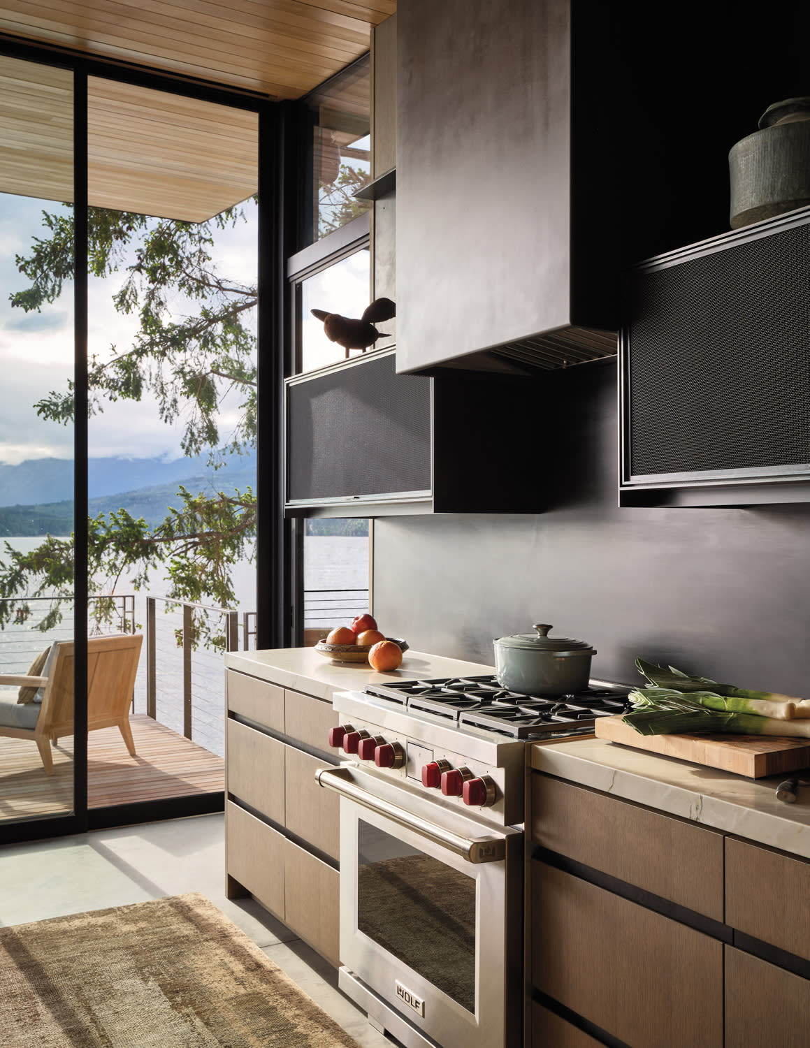 Modern kitchen with a scenic lake view. Features a sleek stove, fresh produce, and an open sliding glass door leading to a wooden deck. Serene and inviting.