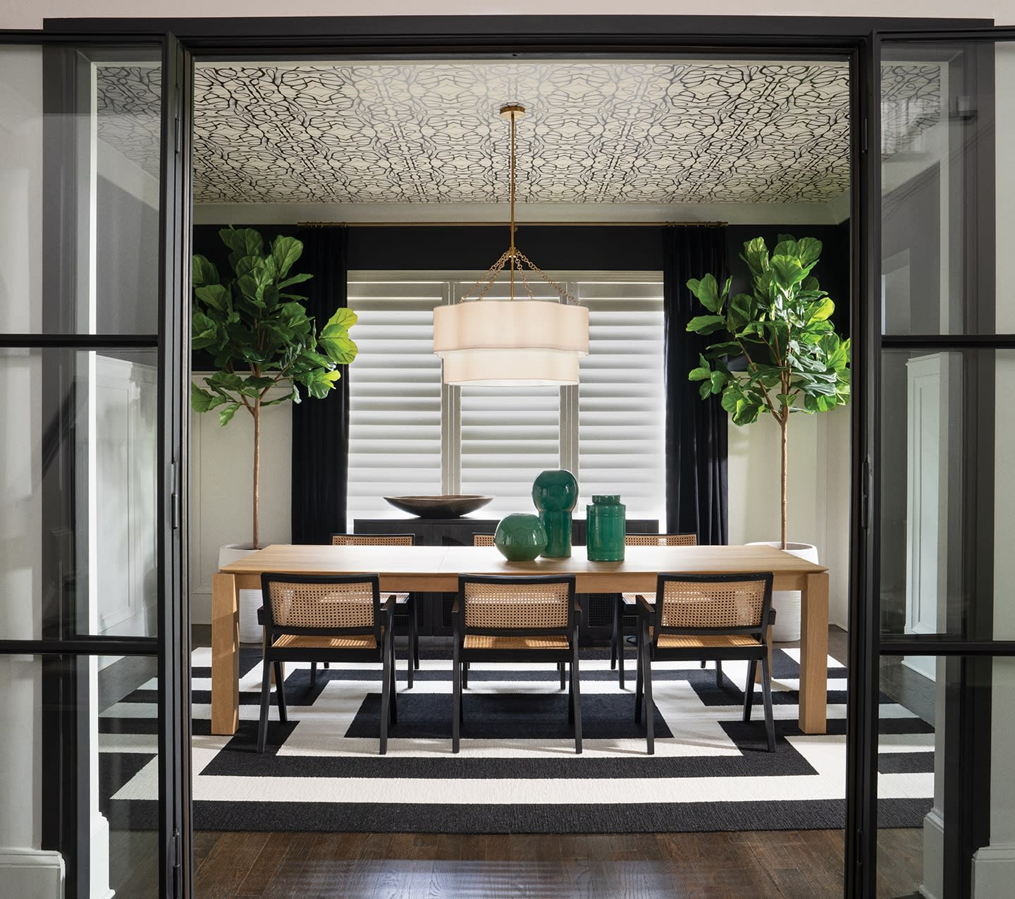 Elegant dining room with a wooden table, six chairs, two large potted plants, black and white rug, modern chandelier, and decorative green vases.