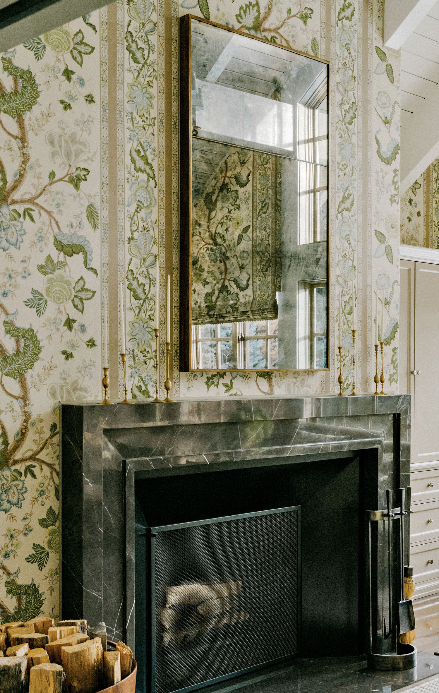 A fireplace with a mirror hanging above the mantel covered in wallpaper with a nature pattern.