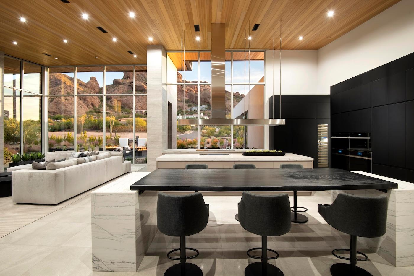 Spacious modern kitchen with large windows showcasing mountain views. Features wooden ceiling, marble island, black chairs, and sleek appliances.