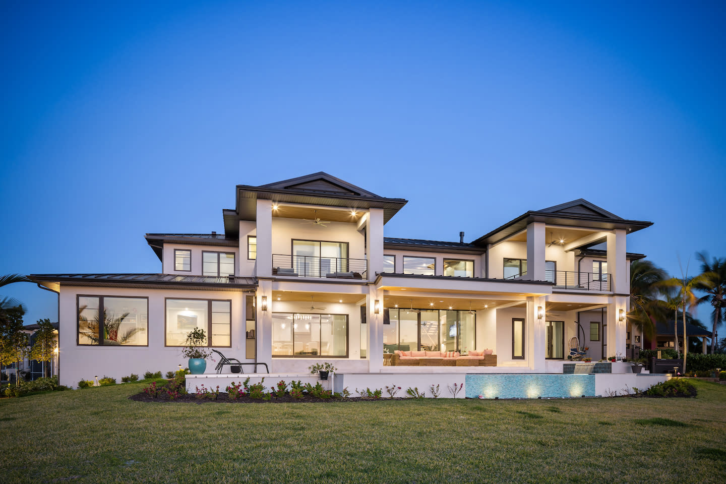 Two-story coastal manor at twilight, its white stucco façade framed by three pavilion-style roof peaks and wide balconies with sleek metal railings. Recessed lights glow beneath deep overhangs, illuminating walls of glass that open to covered terraces, a pink outdoor seating set, and a narrow spill-edge pool cascading into a tiled water feature. A freshly manicured lawn and low flower beds complete the serene evening scene.