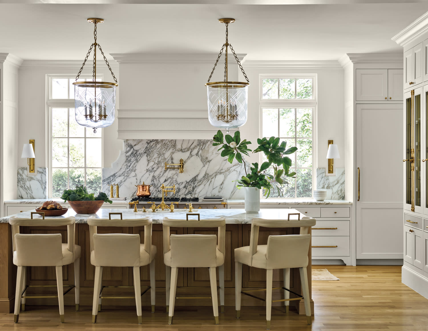 Elegant kitchen with white marble countertops and backsplash, wooden island, plush beige chairs, gold fixtures, and large pendant lights. Bright and airy.