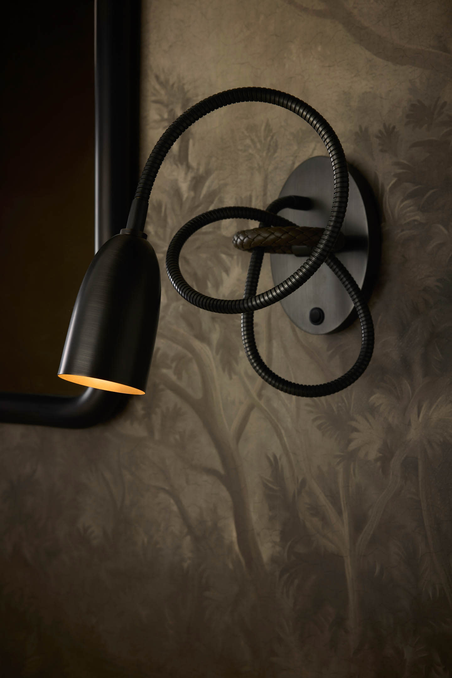 A sleek black wall sconce with a flexible, looped arm emits soft light. The background features a dark floral wallpaper, creating an elegant atmosphere.