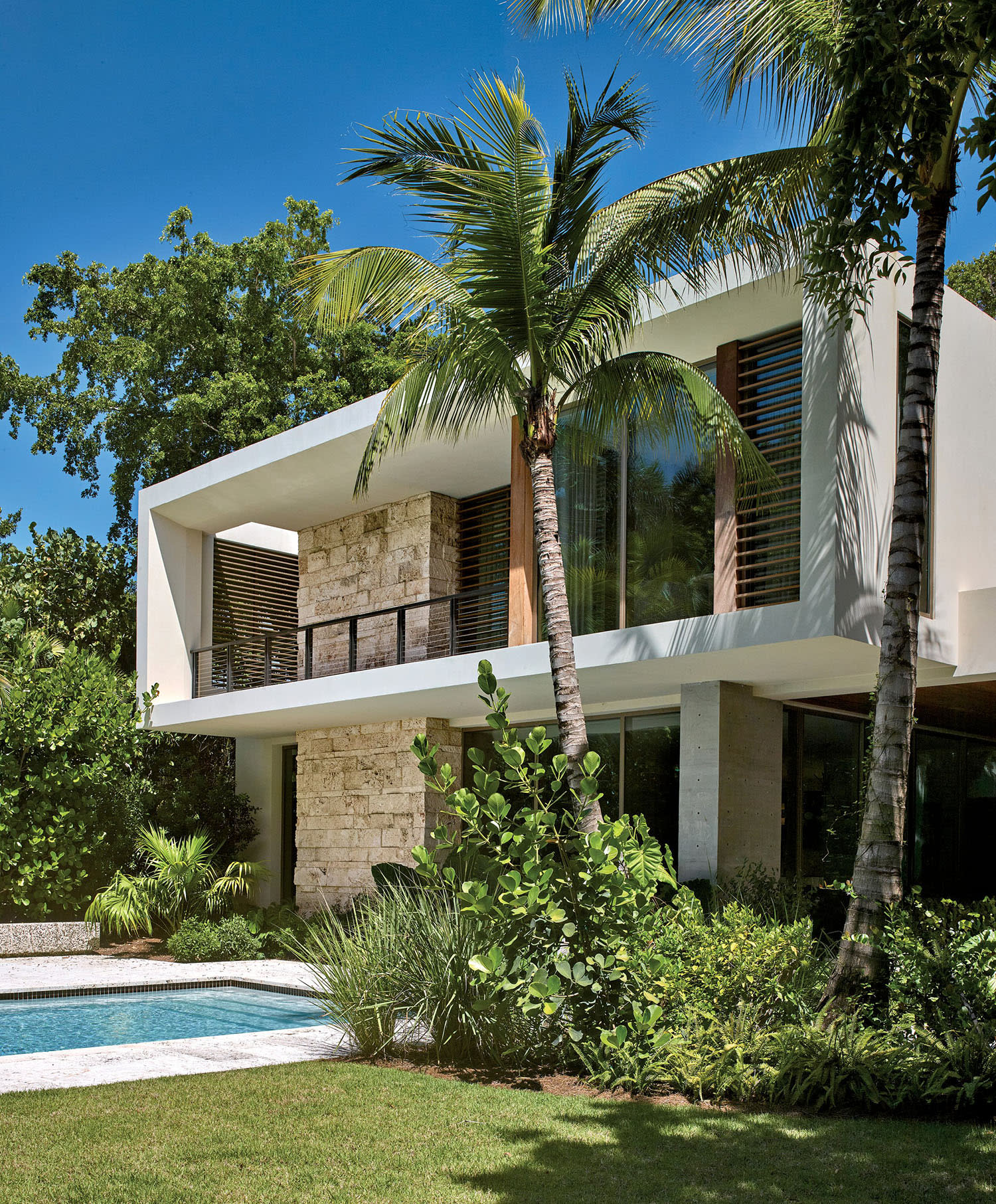 Modern two-story house with stone and wood accents, surrounded by lush greenery and palm trees.