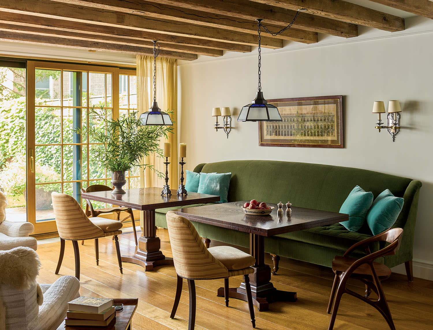Cozy dining area with wooden beams and large windows. Features a green sofa, wooden tables, tan chairs, and blue-green pillows. Inviting and warm ambiance.
