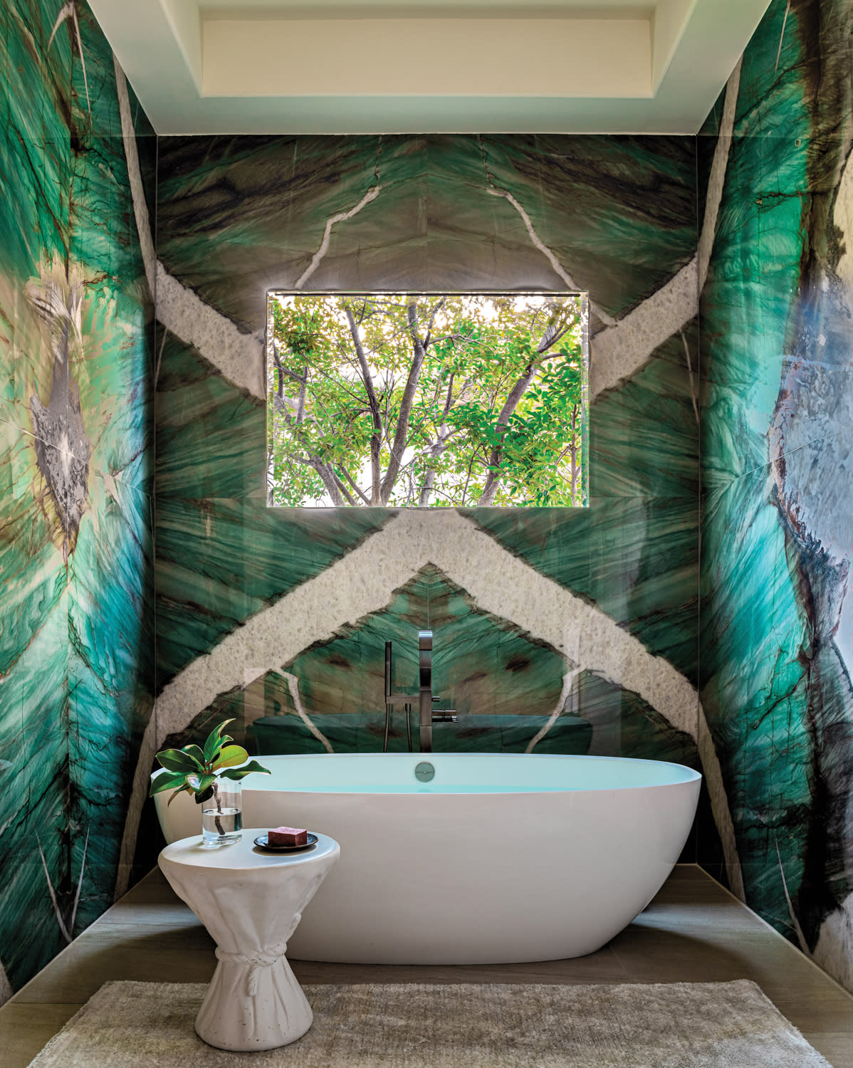 Luxurious bathroom with vibrant green marble walls and a large window framing lush trees. A sleek white bathtub and elegant side table add a modern touch.