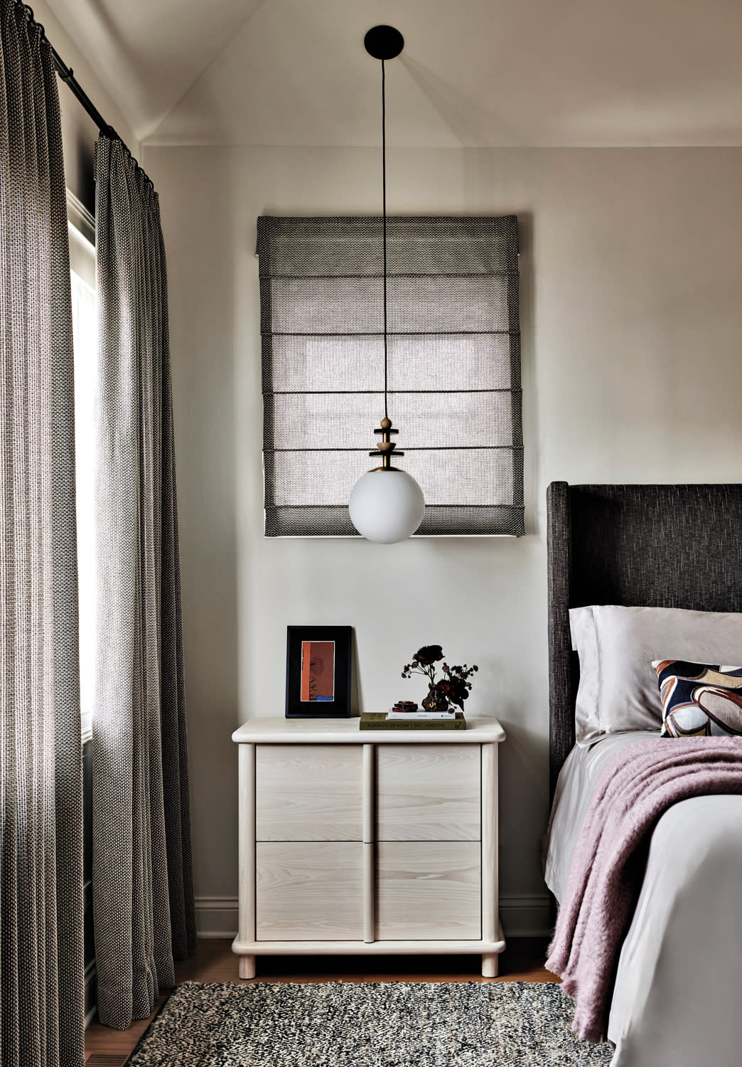 Cozy bedroom with a modern nightstand beside a tall bed. A pendant light hangs above. Curtains and a textured rug add warmth, enhancing the inviting tone.